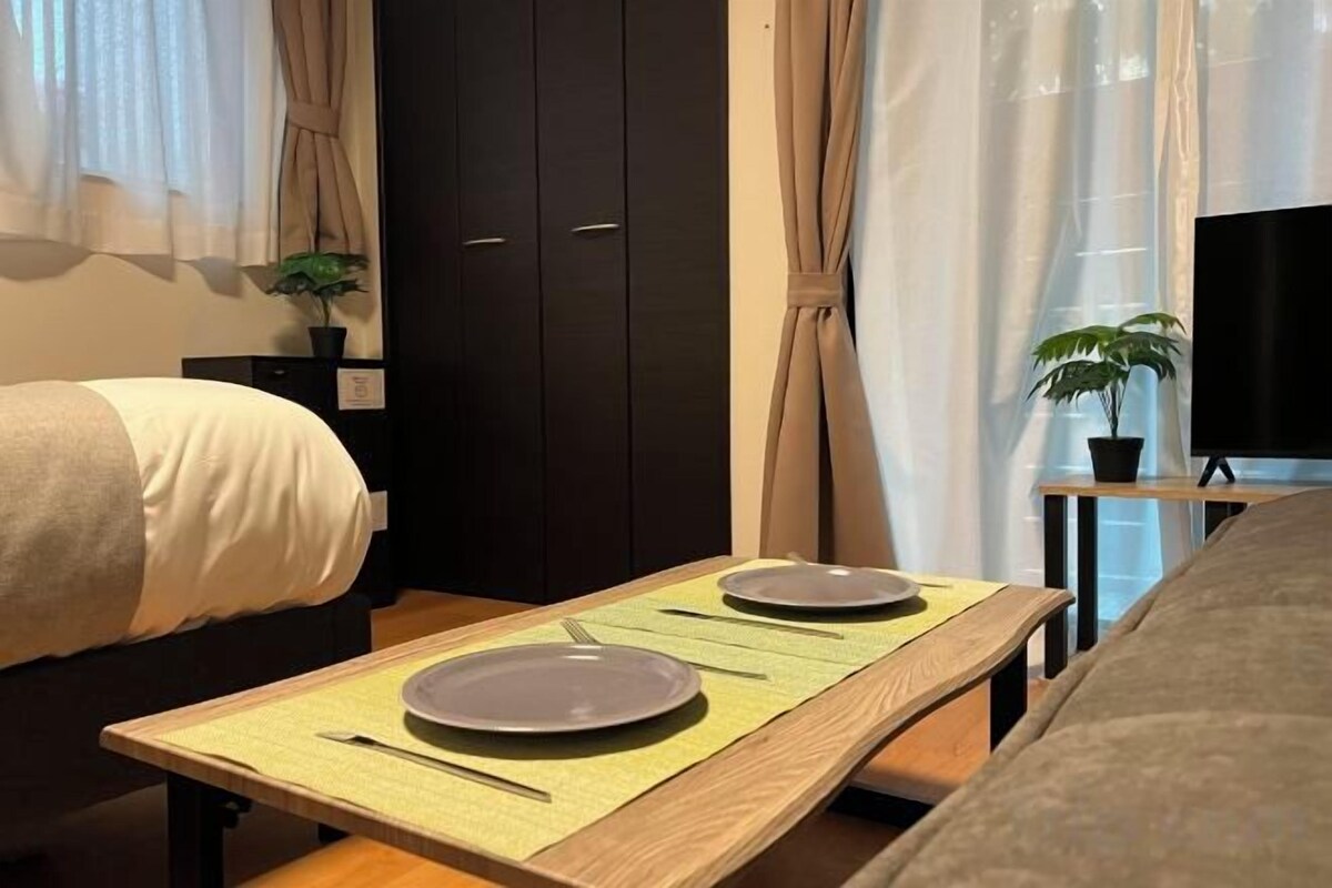 Hotellihuone, jossa on sänky, yöpöytä, jossa on lautanen ja ruokailuvälineet, pieni pöytä, jossa on alusta, ruukkukasvi ja televisio jalustalla.