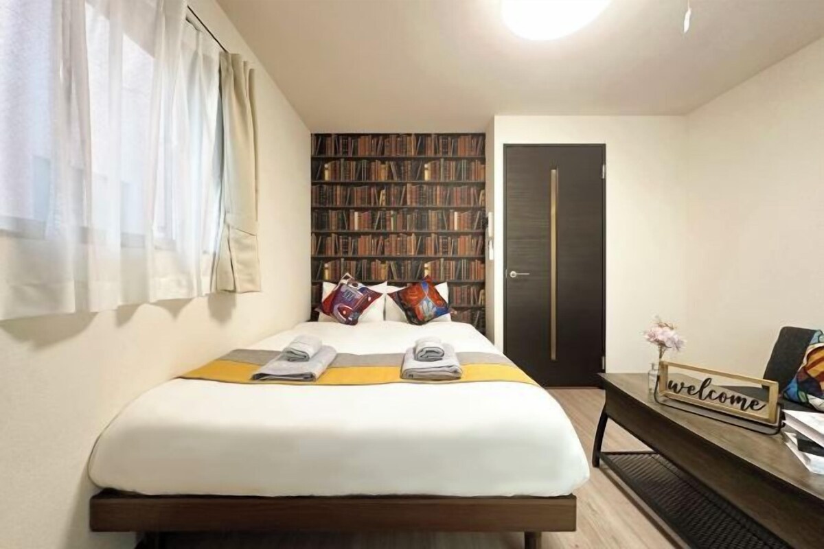 Een net opgemaakt bed met handdoeken, een boekenplank, een 'welcome'-bord en een deur in een hotelkamer.