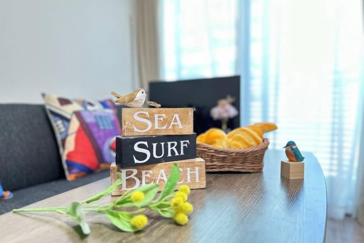 Puinen pöytä, jonka päällä on puisia palikoita, joista on muodostettu teksti ”SEA SURF BEACH”. Pöydällä on myös lintuhahmo, kori appelsiineja ja pieni puinen lintuhahmo.