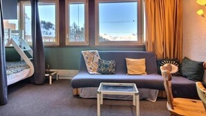 A cozy room with a wooden ceiling, a sofa, a small table, and a view of a snowy landscape through large windows. - Spacious studio with wifi in Plagne Centre, near slopes (La Plagne)