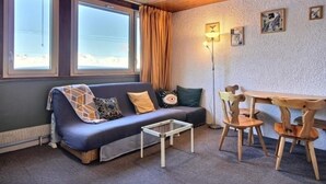 A cozy room with a blue sofa, a small table, and a wooden chair. There are windows with curtains, a lamp, and a wall with framed pictures. - Spacious studio with wifi in Plagne Centre, near slopes (La Plagne)