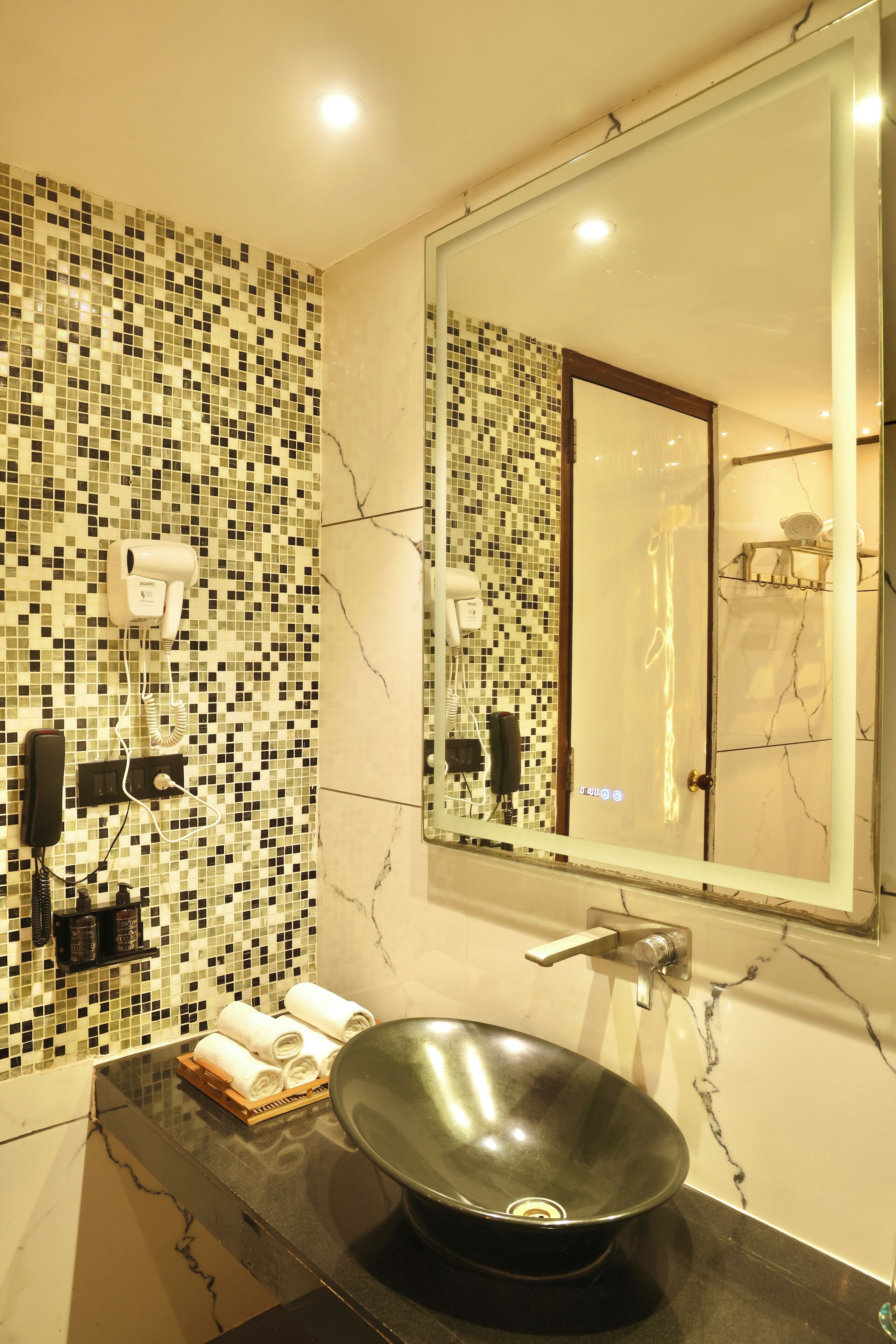 A bathroom with a mosaic tile wall, a large mirror, a black sink, and a towel rack with towels.
