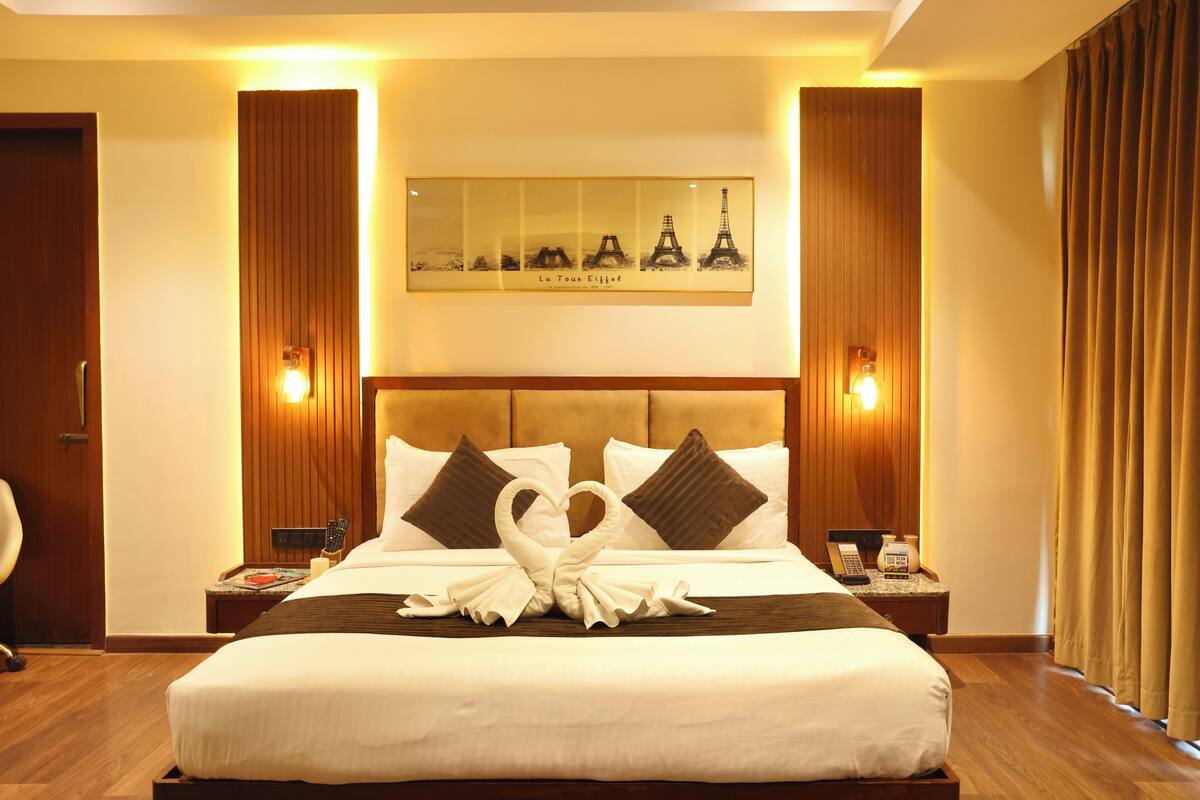 A hotel room with a large bed, a headboard with a mirror, two bedside tables, and a wall-mounted artwork of the Eiffel Tower.