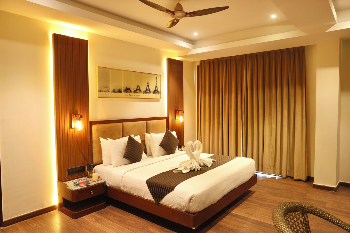 A hotel room with a large bed, wooden headboard, wall-mounted lights, a ceiling fan, and a view of the Eiffel Tower through a window.
