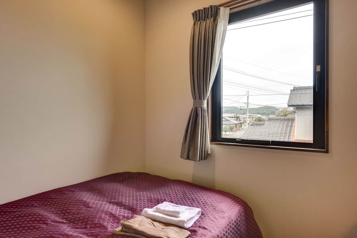 Ein Einzelbett mit einem gemusterten Überwurf, ein Fenster mit Vorhängen und Blick auf Gebäude.
