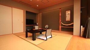 Ein traditioneller japanischer Raum mit Tatami-Boden, einem niedrigen Tisch mit Kissen, einem Fernseher und einem Regal mit Dekorationen.