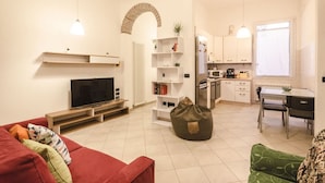 A modern living room with a flat-screen TV, a red sofa, a green bean bag, and a kitchen area with white cabinets and a dining table. - Appartamento Schiavonia (Bologna)