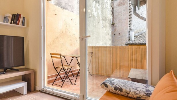 A room with a sliding glass door leading to an outdoor area with a wooden table and chairs, a television on a stand, and a bed with a floral pillow. - Ca' di Leo (Bologna)