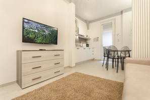 A modern living room with a flat-screen TV, a beige sofa, a dining area with a glass table and chairs, and a kitchen in the background.