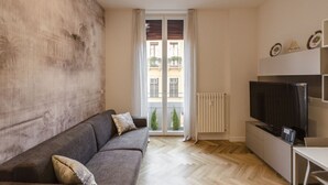 A modern living room with a grey sofa, a flat-screen TV, and a large window with a view of buildings and greenery.
