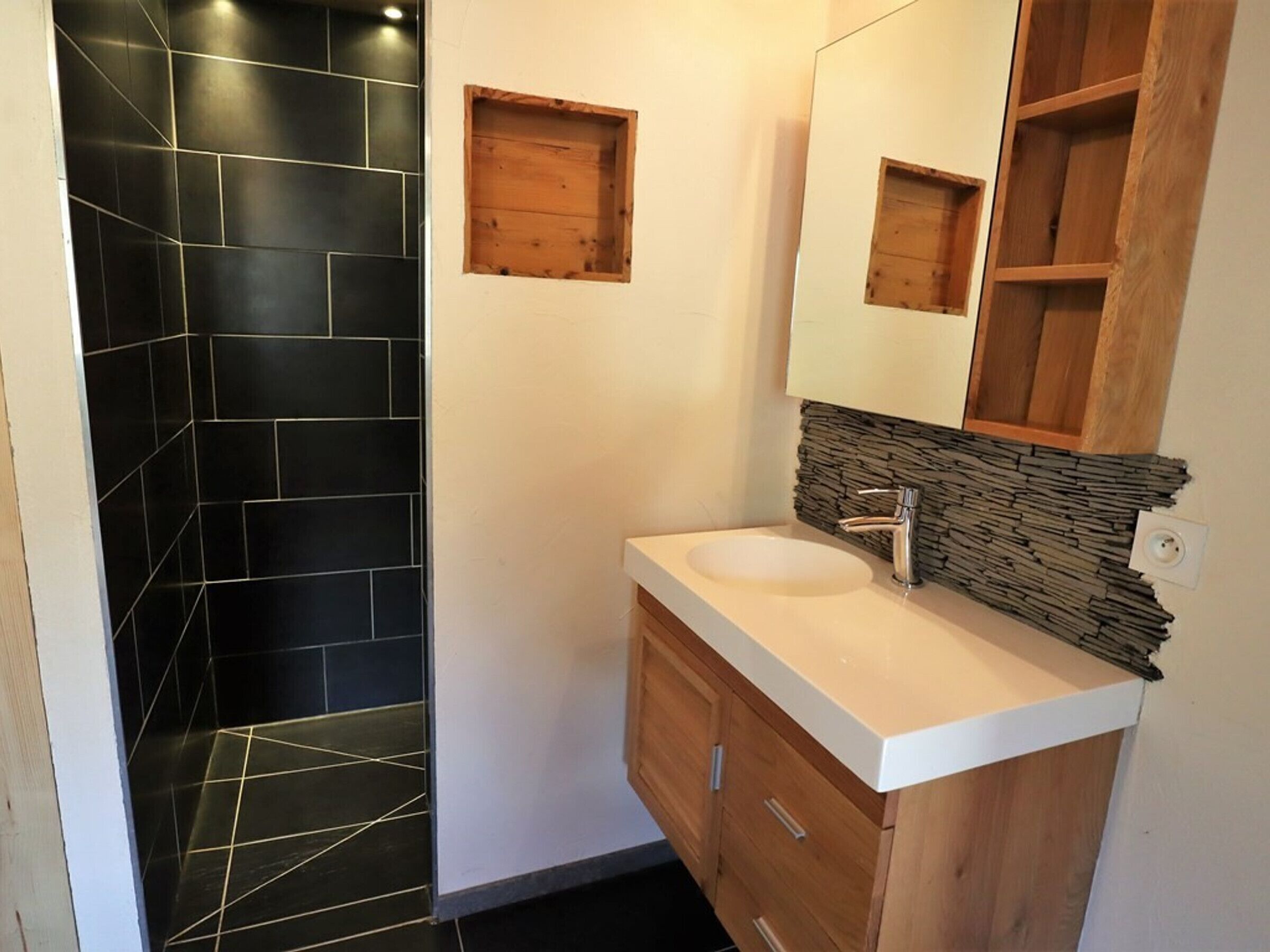 Een badkamer met een douche in zwart tegels, een houten wastafelmeubel met een witte wasbak en een accentmuur van steen.