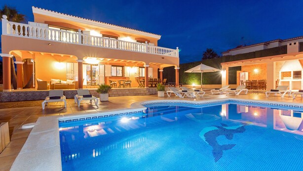 A large outdoor swimming pool with lounge chairs and umbrellas, a covered patio area with seating, and a two-story building with balconies.