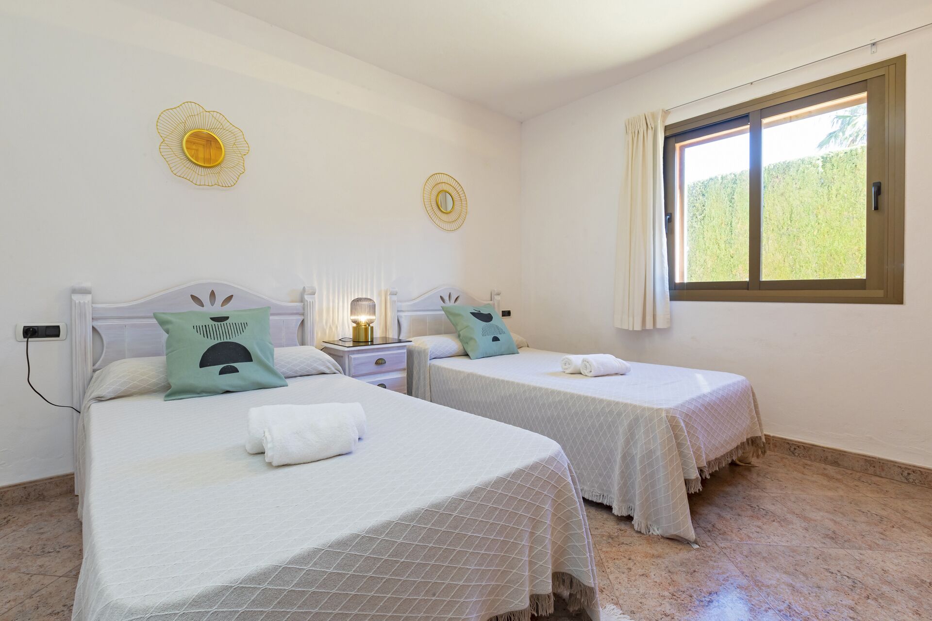 Un dormitorio limpio y bien iluminado con dos camas, cada una con cubrecama y almohadas a juego. Cuenta con una ventana con cortinas, una mesita de noche con lámpara y un adorno en la pared.