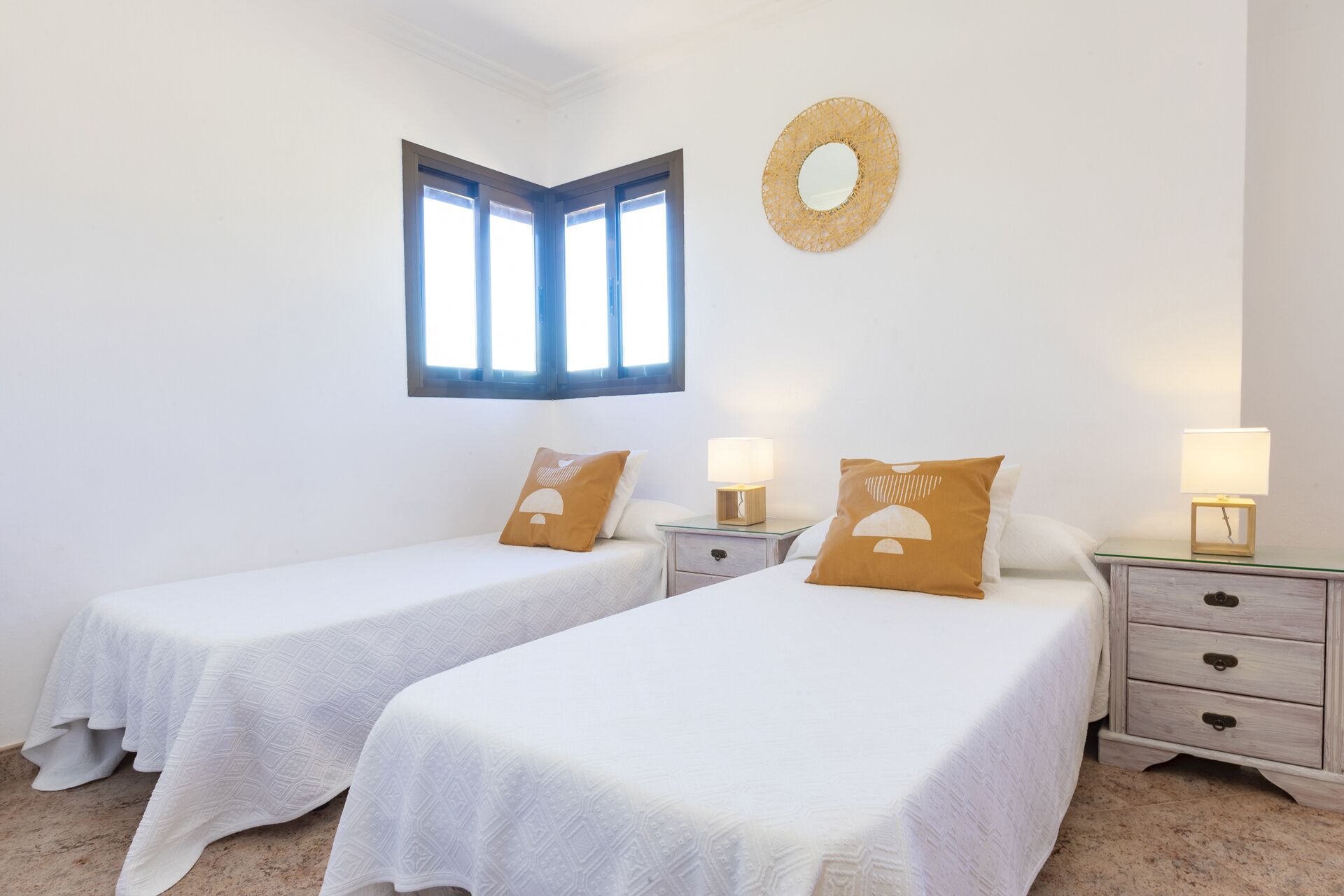 Una habitación con dos camas, una ventana, un espejo montado en la pared y una mesita de noche con lámpara.