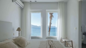 A bedroom with a large bed, two windows with a view of the sea, a bedside table with a lamp, and a bathroom visible through an open door.