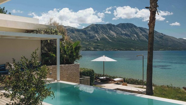 A pool area with lounge chairs and an umbrella, overlooking a lake with mountains in the background.