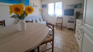 Ein Essbereich mit rundem Tisch, Stühlen, einer Vase mit Sonnenblumen, einem Gemälde, einem Fernseher und einem Fenster mit Vorhängen.