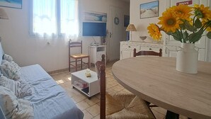 Ein Wohnzimmer mit einer Couch, einem Esstisch mit einer Vase voller Sonnenblumen, einem Fernseher und einer Küchenzeile mit einem 'Drive'-Schild.