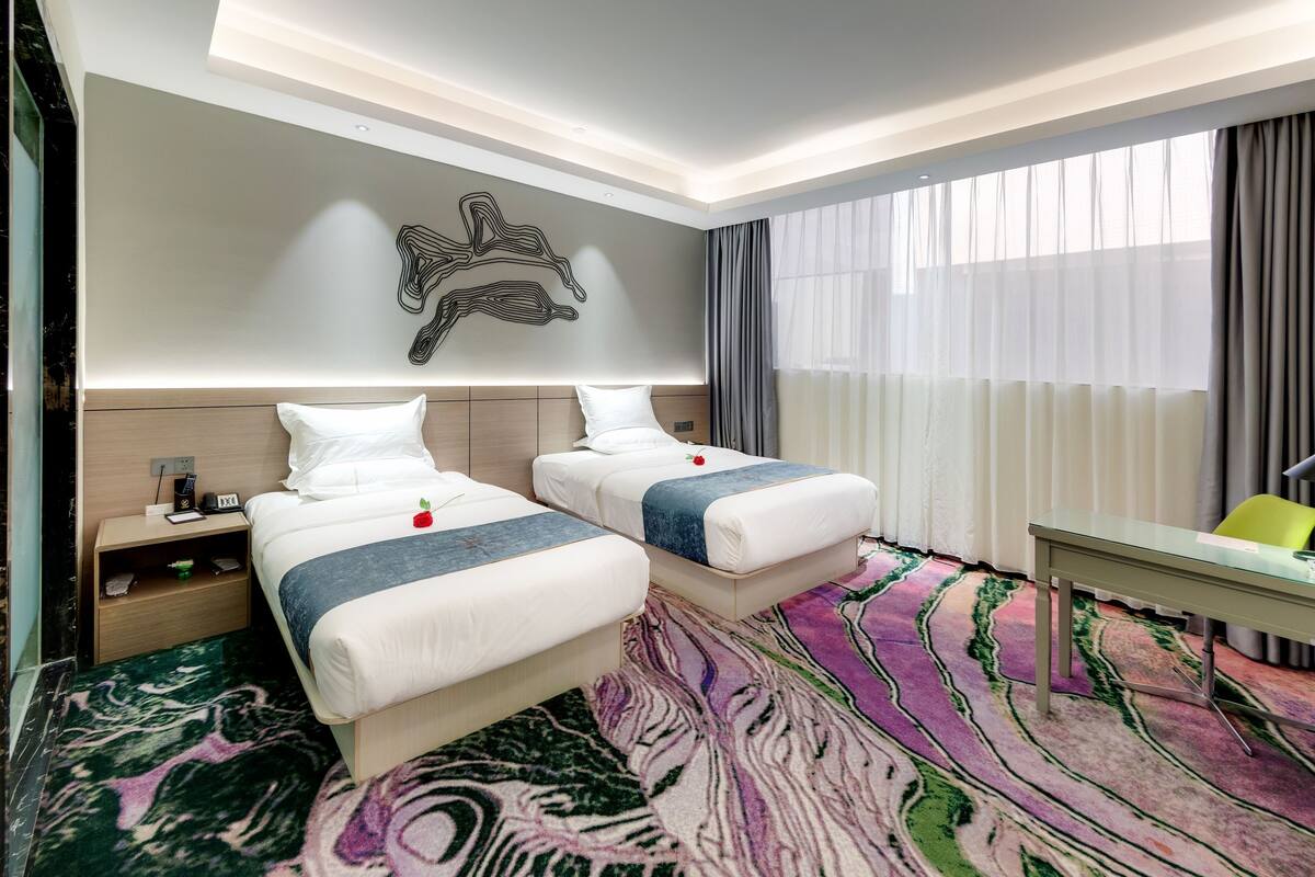 Une chambre d’hôtel avec deux lits, un bureau, une chaise, un élément décoratif mural et une grande fenêtre avec des rideaux.