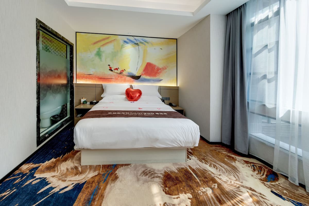 Une chambre d’hôtel avec un grand lit, une peinture abstraite aux couleurs vives au-dessus du lit, une décoration en forme de cœur rouge, une tête de lit en bois, une table de chevet, un téléphone, une fenêtre avec des rideaux légers et une moquette à motifs.