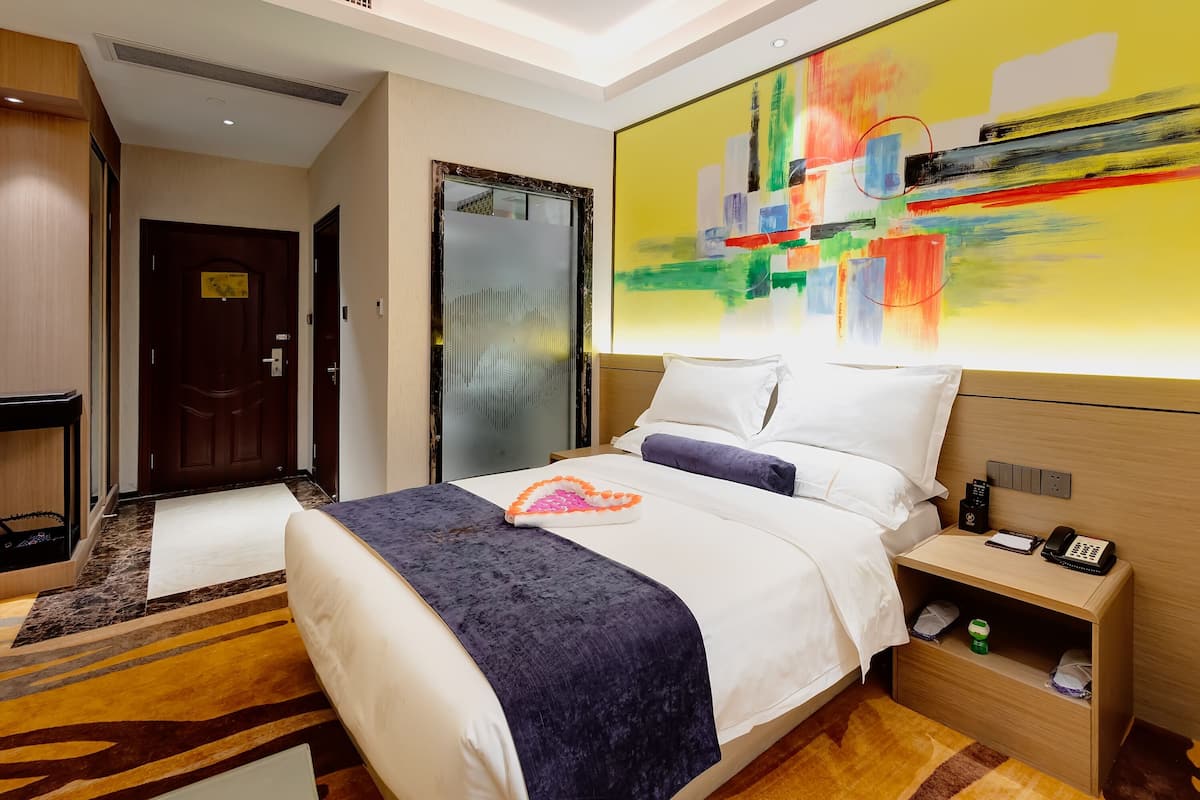 Une chambre d’hôtel avec un grand lit, une table de chevet avec un téléphone et un bloc-notes, une commode et une peinture abstraite aux couleurs vives accrochée au mur.
