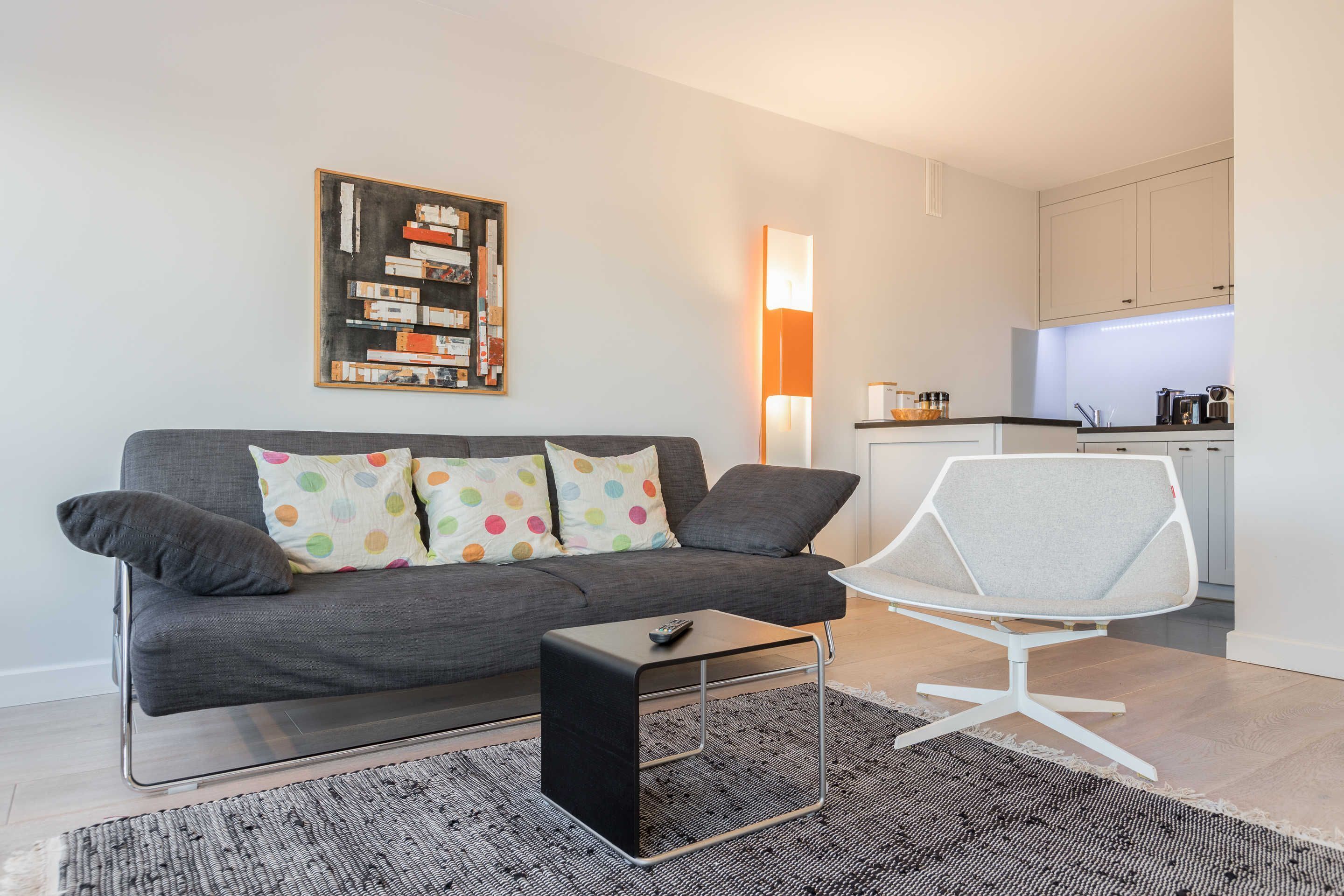 A modern living room with a grey sofa, a white chair, a coffee table, and a kitchen in the background.
