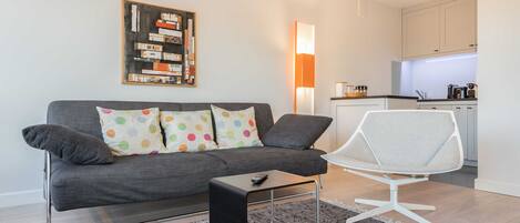 A modern living room with a grey sofa, a white chair, a coffee table, and a kitchen in the background.