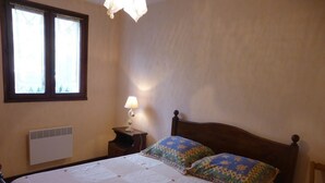 Ein Schlafzimmer mit einem hölzernen Bett, zwei Kissen, einem Nachttisch mit Lampe, einem Fenster mit Vorhängen und einem Heizkörper.