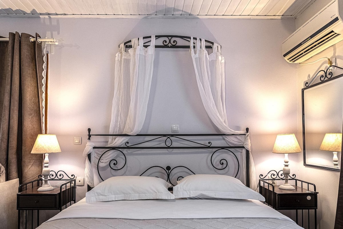 A bedroom with a four-poster bed, two bedside tables with lamps, a ceiling fan, and an air conditioning unit.