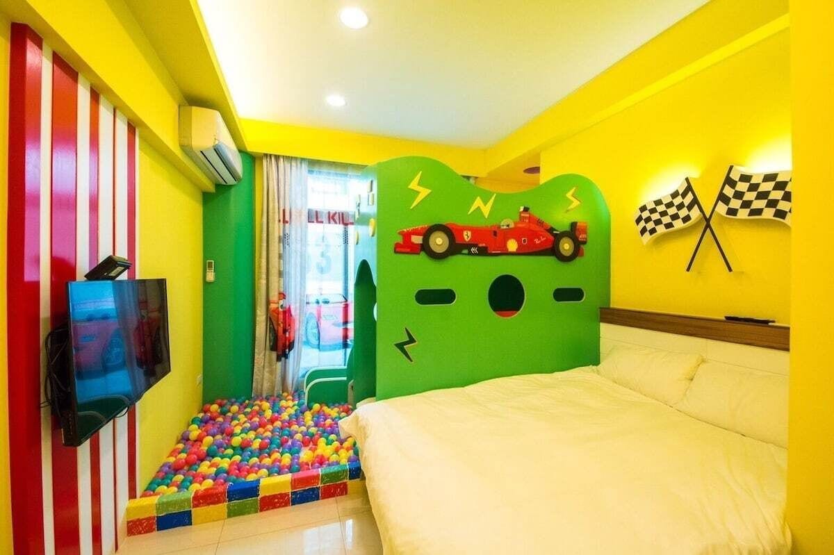 A room with a bed, a TV, a colorful ball pit, and a wall with a race car and flags.