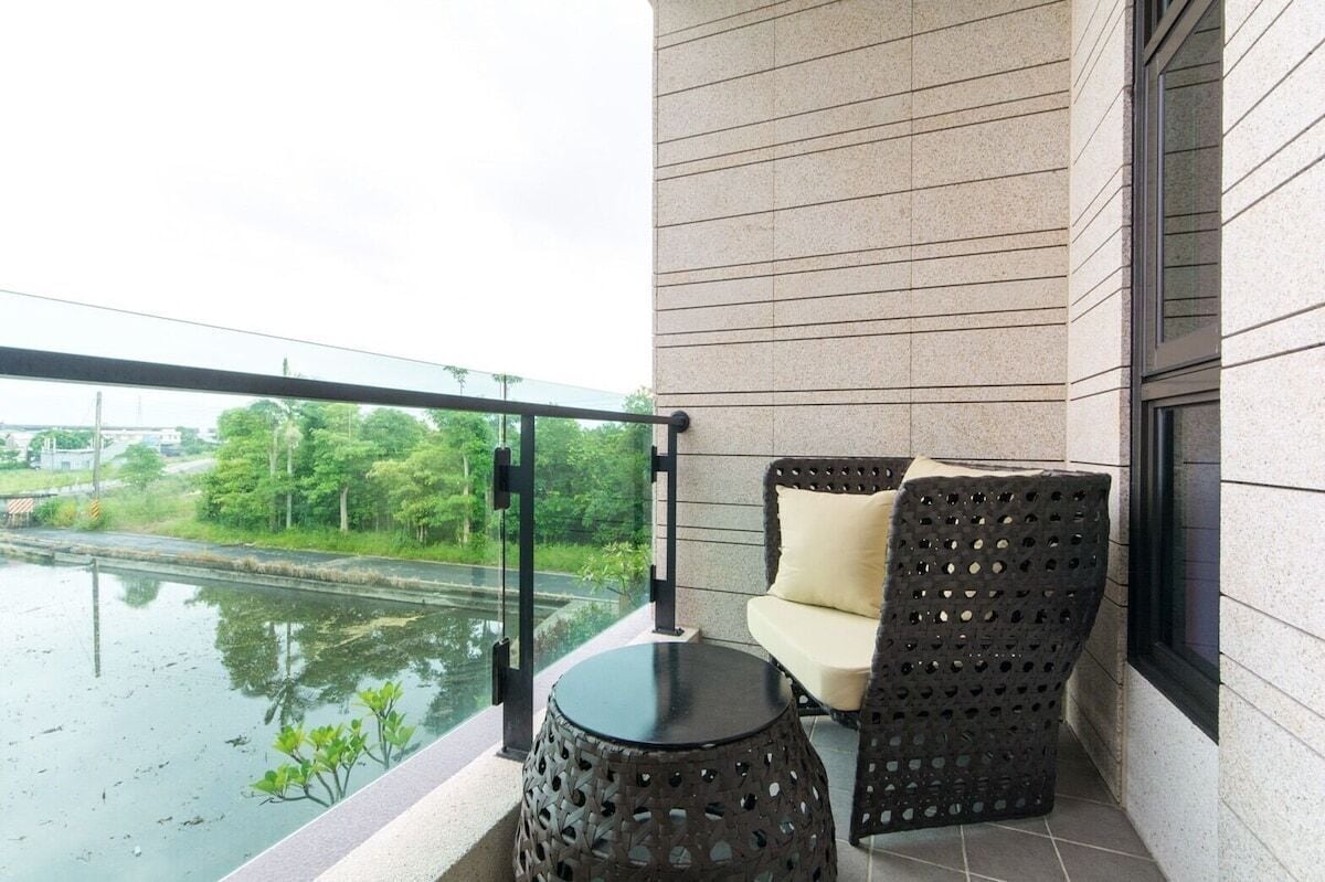 A balcony with a view of a river, a black woven chair, a small round table, and a glass railing.