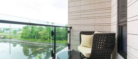 A balcony with a view of a river, a black woven chair, a small round table, and a glass railing.