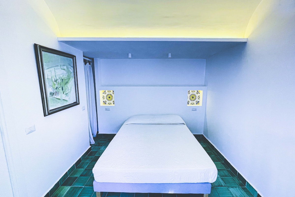 A small, narrow room with a bed, a framed picture on the wall, and two wall-mounted light switches.