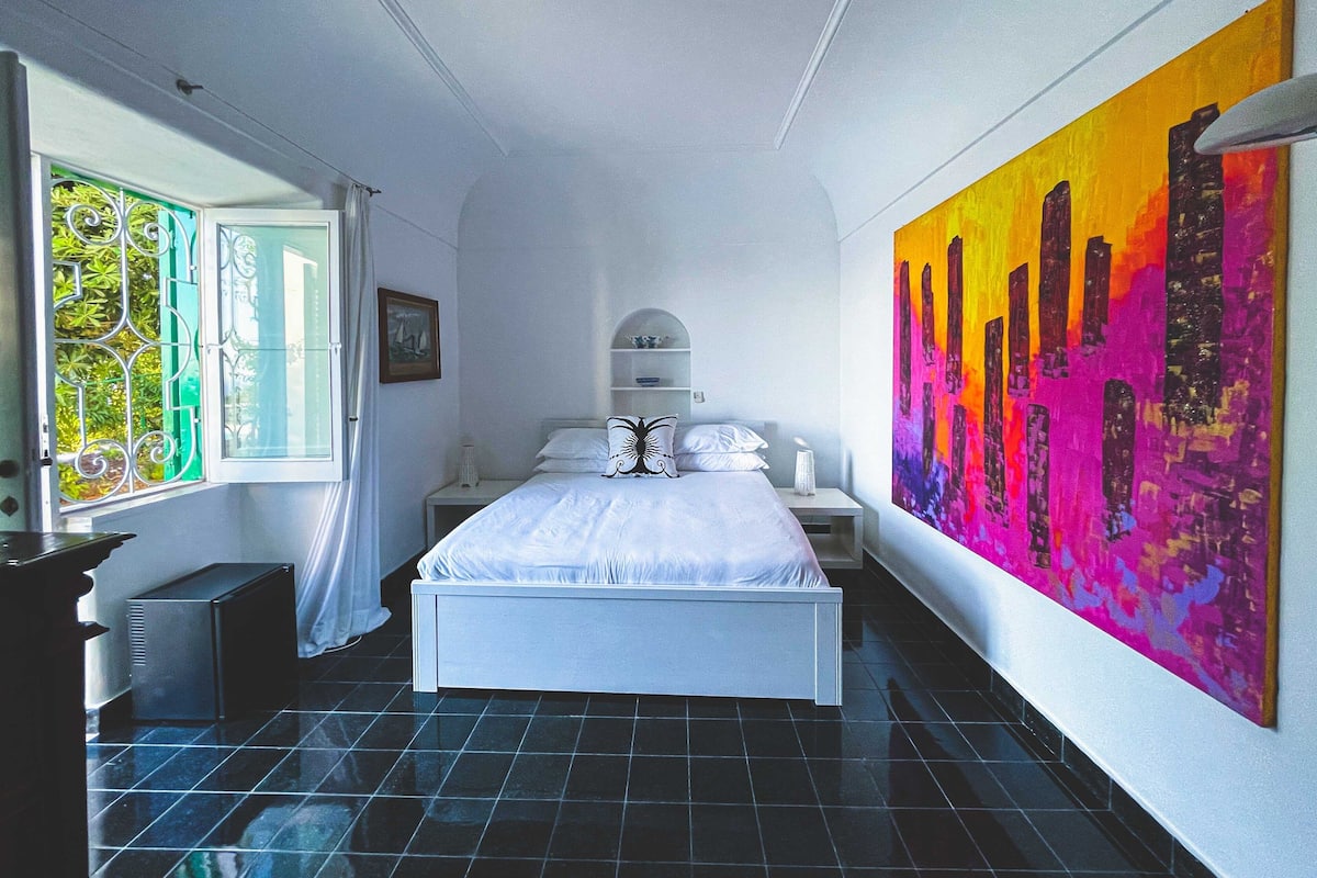 A bedroom with a bed, a nightstand, a window with curtains, and a large colorful painting on the wall.