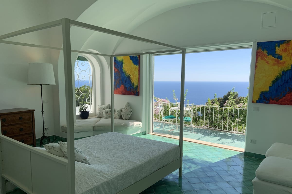 A bedroom with a four-poster bed, a sofa, and a balcony with a view of the sea.