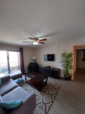 A living room with a sofa, a coffee table, a TV, a ceiling fan, a potted plant, and a sliding door leading to a balcony.