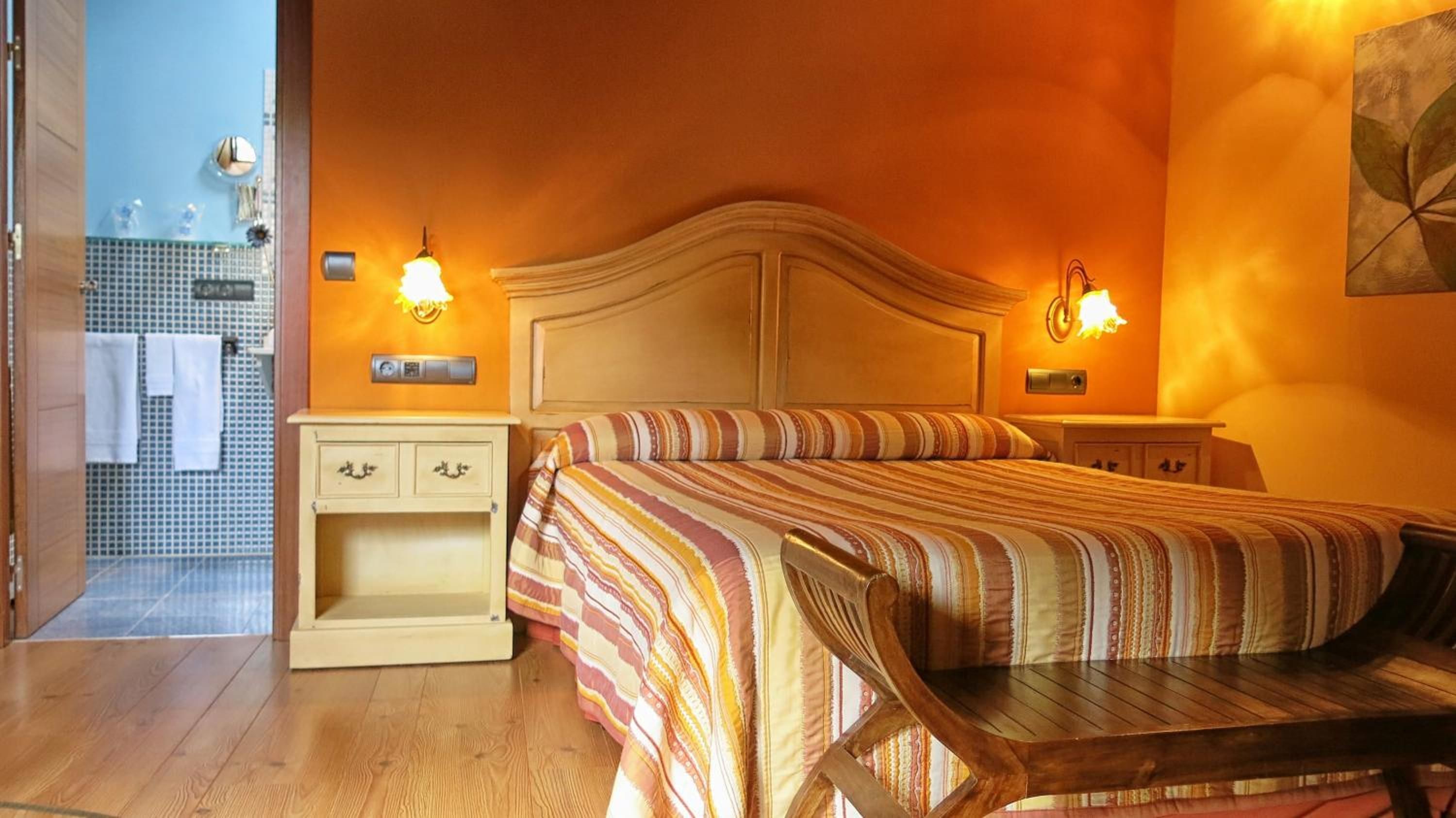 Habitación de hotel con cama, mesitas de noche, una silla de madera y un baño visible a través de una puerta abierta.