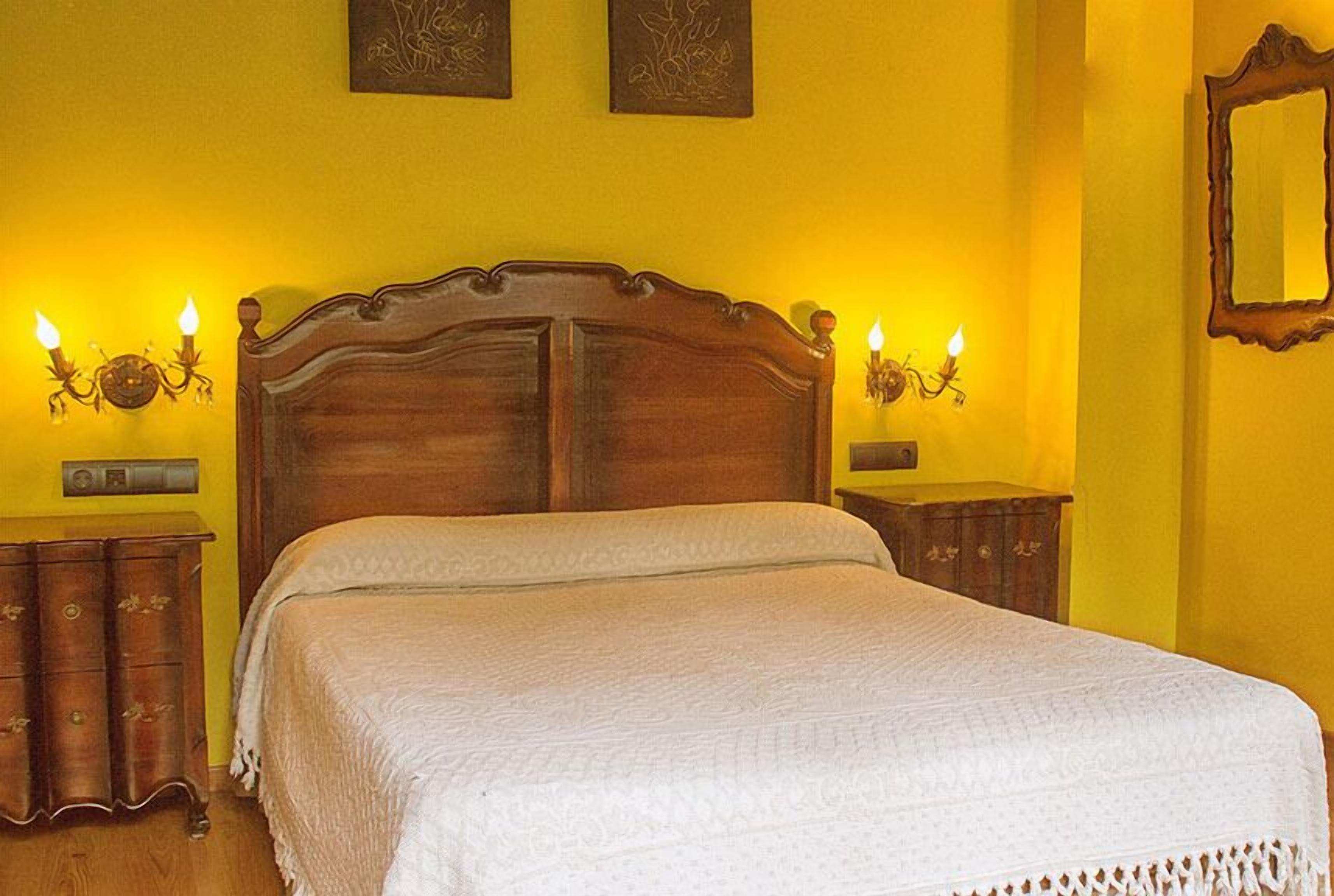 A bedroom with a wooden bed, two wall-mounted sconces, a bedside table, and a mirror.