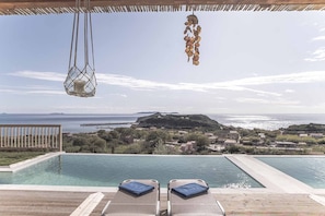 Une vue sur la piscine, avec deux chaises longues, un luminaire suspendu et une vue sur la mer et le paysage lointain.