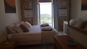 Une chambre à coucher avec un lit, des oreillers, une table de chevet, une lampe, une fenêtre avec des rideaux et une vue sur la verdure à l’extérieur.