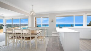 Un comedor amplio con vista al mar, mobiliario blanco y una cocina moderna con ventanales grandes.