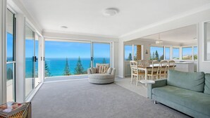 Un salón amplio con un gran ventanal con vistas al mar, un comedor con mesa y sillas de madera, y un sofá verde.