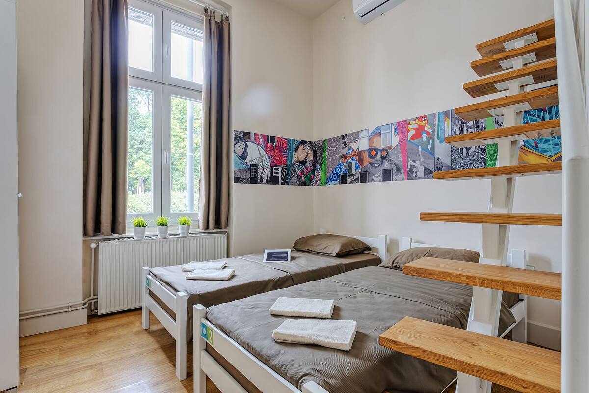 A bedroom with two beds, a window with curtains, a radiator, and a wall with colorful artwork.
