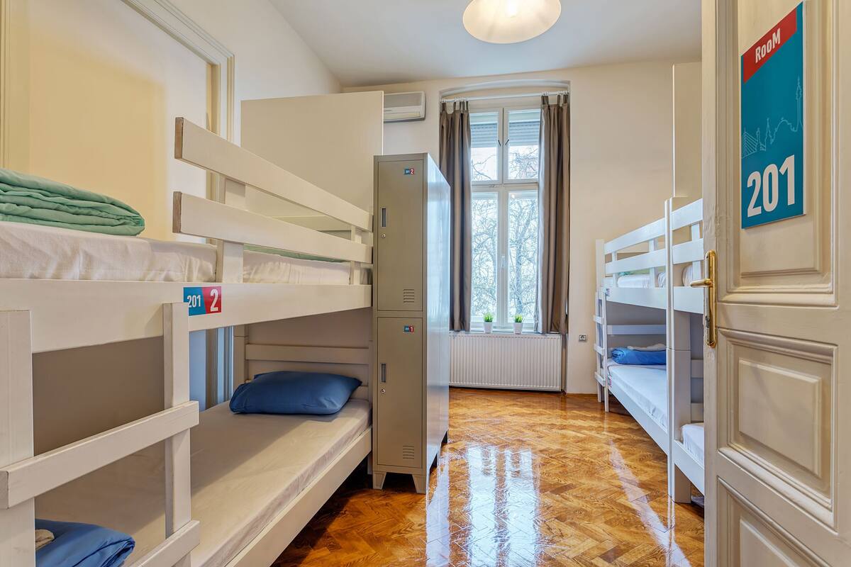 A hotel room with bunk beds, a radiator, a window with curtains, and a door with a room number sign.