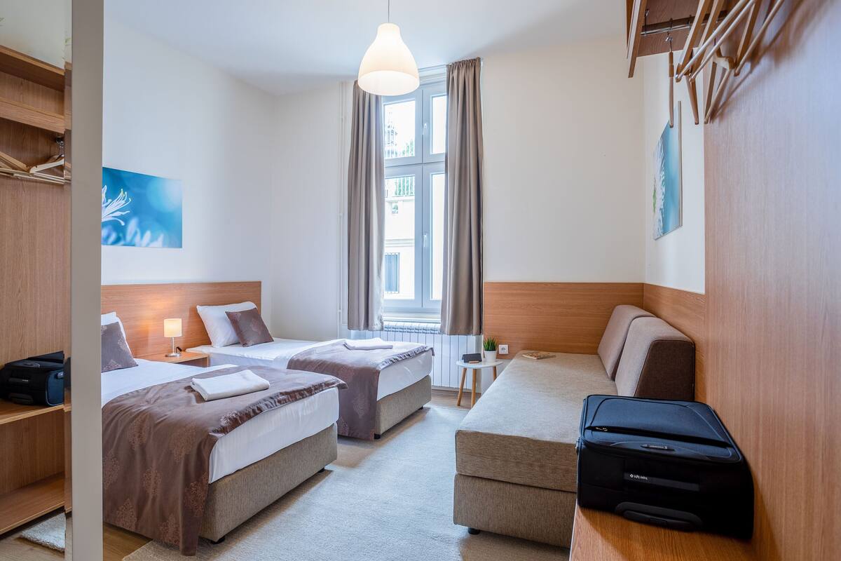 A hotel room with two beds, a sofa, a desk, a suitcase, and a window with curtains.