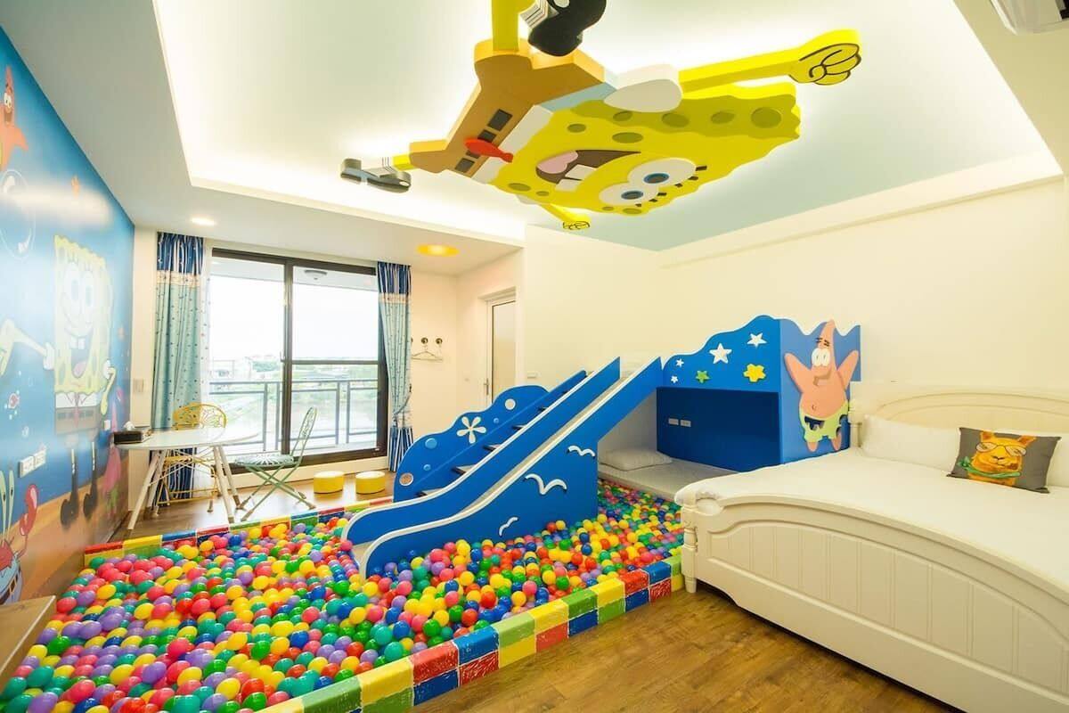 A children's play area with a ball pit and a slide, a bed with cartoon-themed headboard, and a window with curtains.
