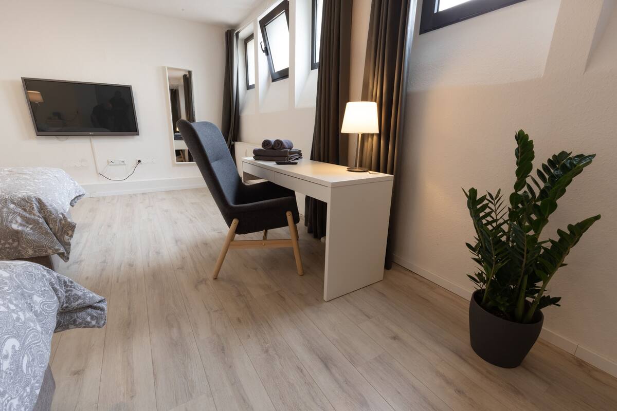 A hotel room with a bed, a desk, a chair, a TV, a lamp, and a potted plant.