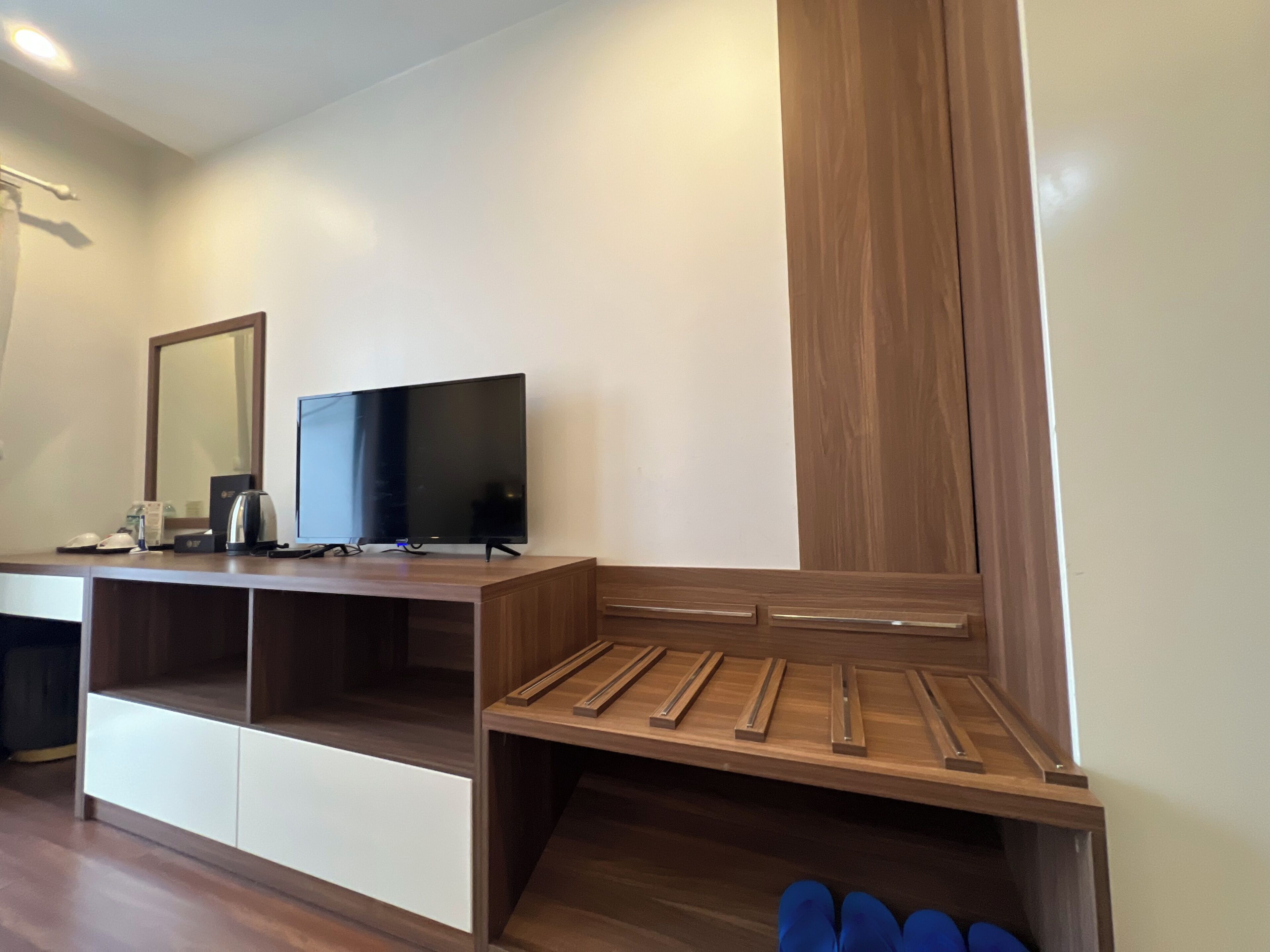 A hotel room with a flat-screen TV on a wooden stand, a mirror, and a wooden bench with storage compartments.