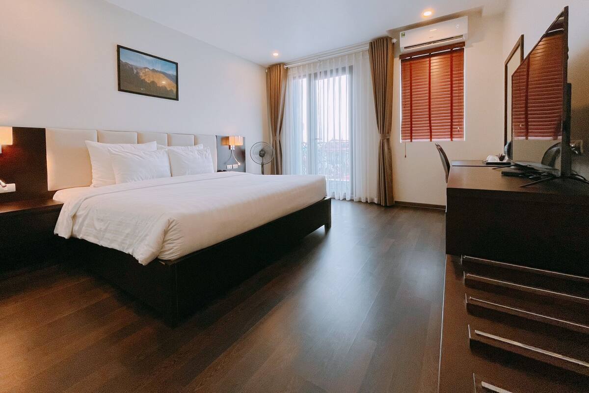 Habitación de hotel con una cama grande, un escritorio, una silla, una lámpara de escritorio, un ventilador de pie y una ventana con cortinas.