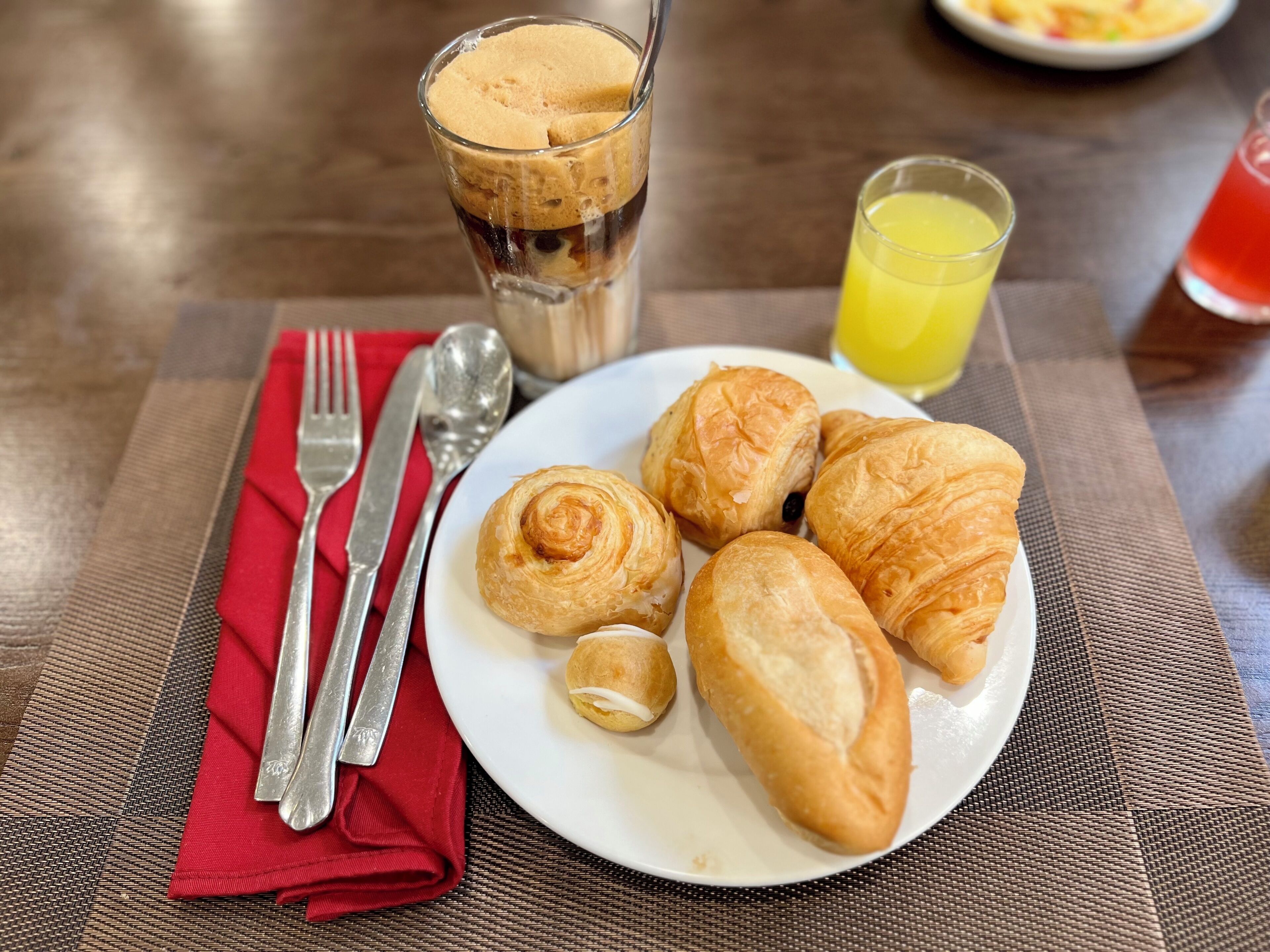 A plate of croissants, a glass of iced coffee, and a glass of orange juice on a table with a red napkin and cutlery.